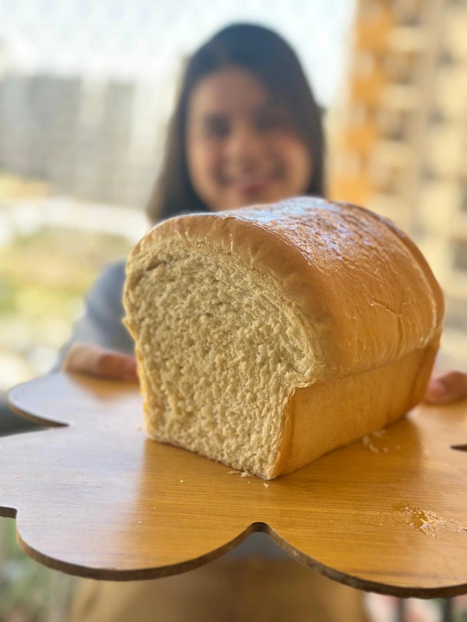 Pão caseiro em primeiro plano, com a Mari sorrindo ao fundo